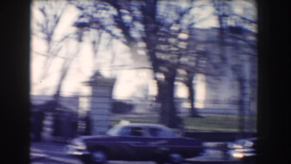 Passing cars cross the foreground of a 1960 Washington DC street view with bare trees, lamps, and a white building beyond.