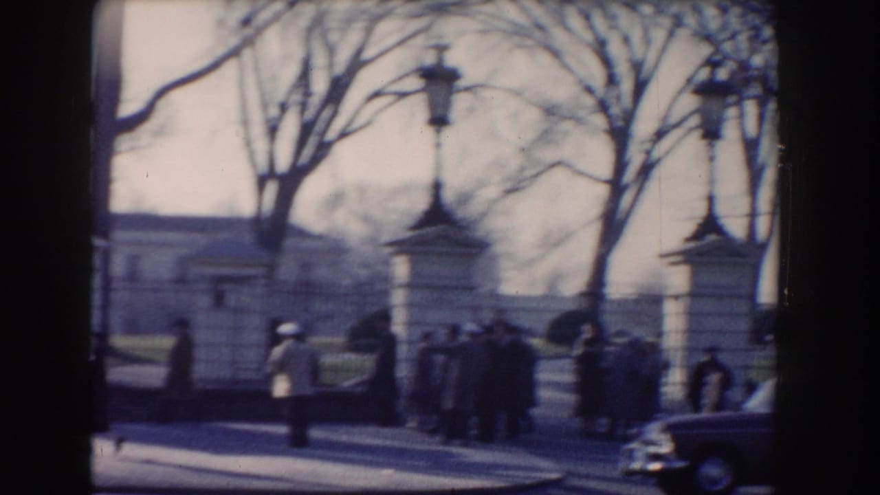 Pedestrians near gate posts, bare trees, and a dark car outside a formal grounds entrance.