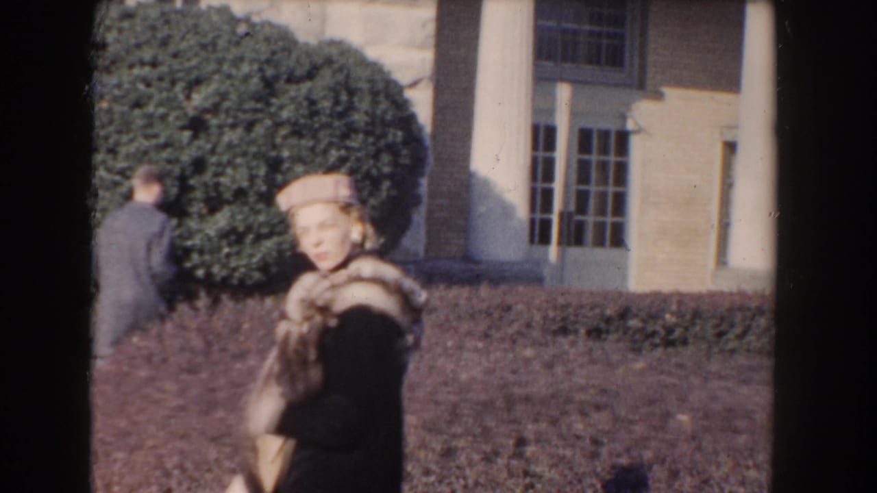 Woman in a hat and fur stole stands near a large hedge and columned stone building.