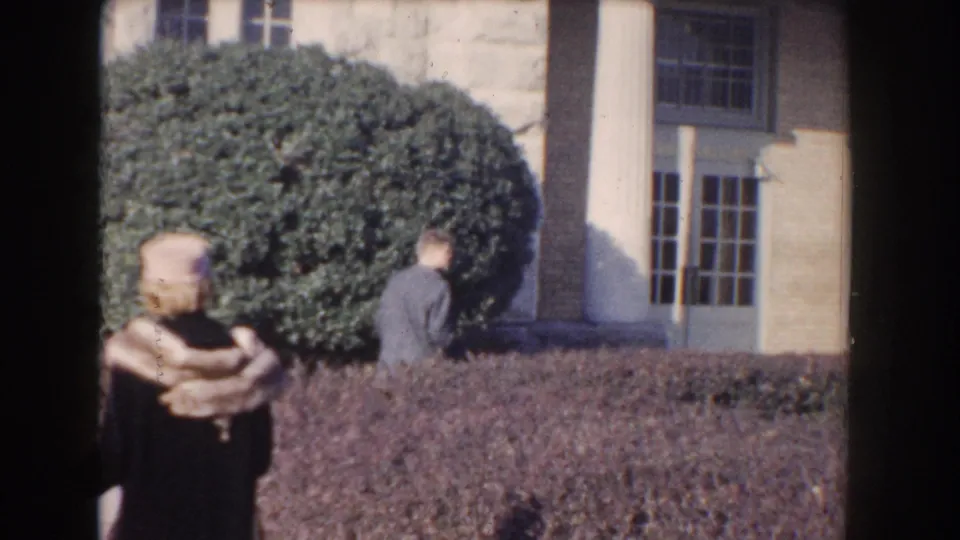 1960 Washington DC home-movie frame of a woman in a hat and fur stole near a hedge, with a man and columned facade behind her.