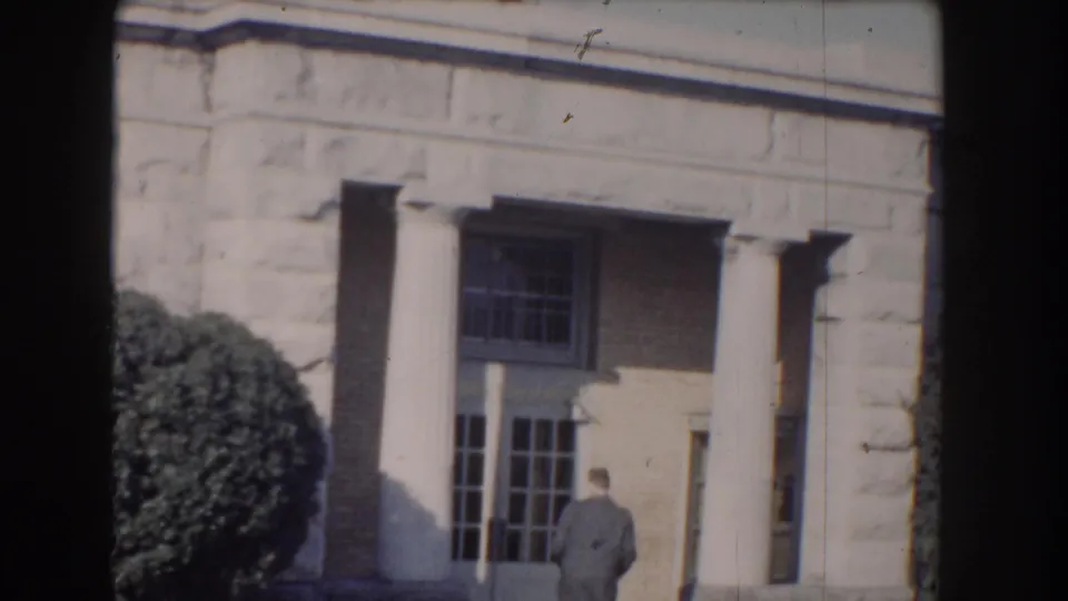 1960 Washington DC frame showing a formal stone entrance with columns, windows, hedge, and a man walking near the doorway.