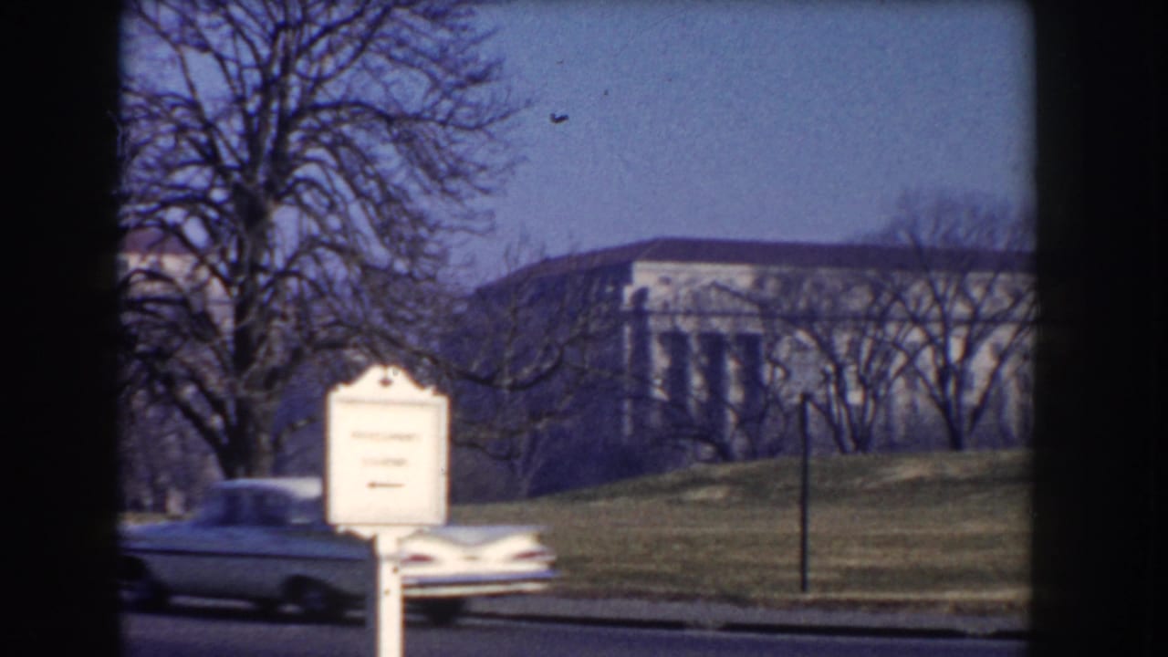 A classic American car passes a roadside sign in Washington DC, with formal public buildings and winter trees in the background.