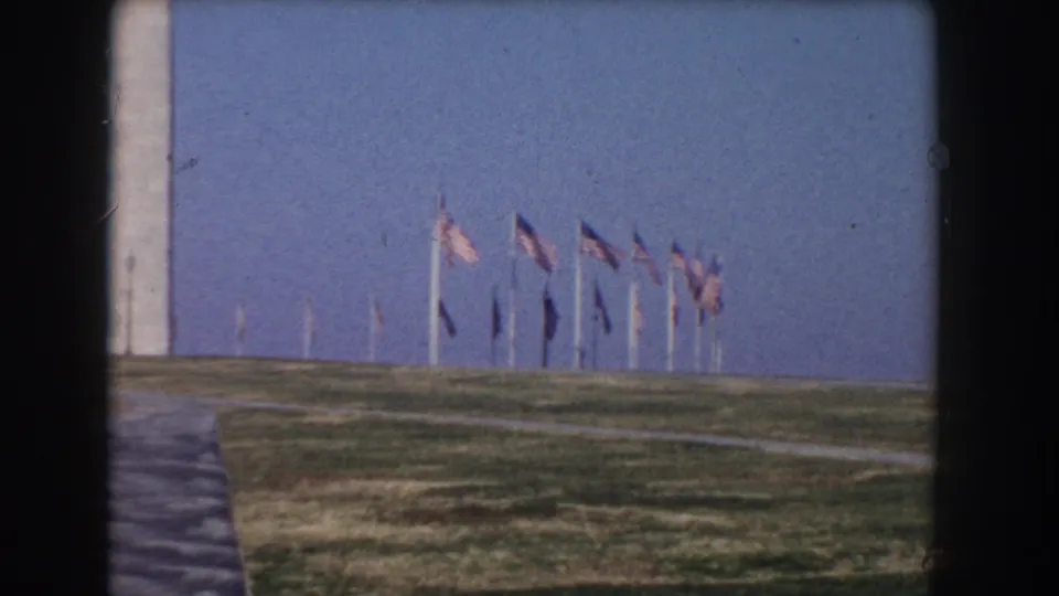 Washington Monument grounds with rows of American flags, walkways, open lawn, and blue sky in a 1960 8mm home-movie frame.