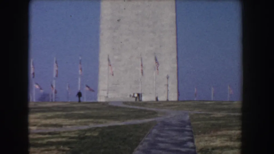 Distant visitors walk near the Washington Monument base as flags line the grounds in Washington DC, filmed on 8mm in 1960.