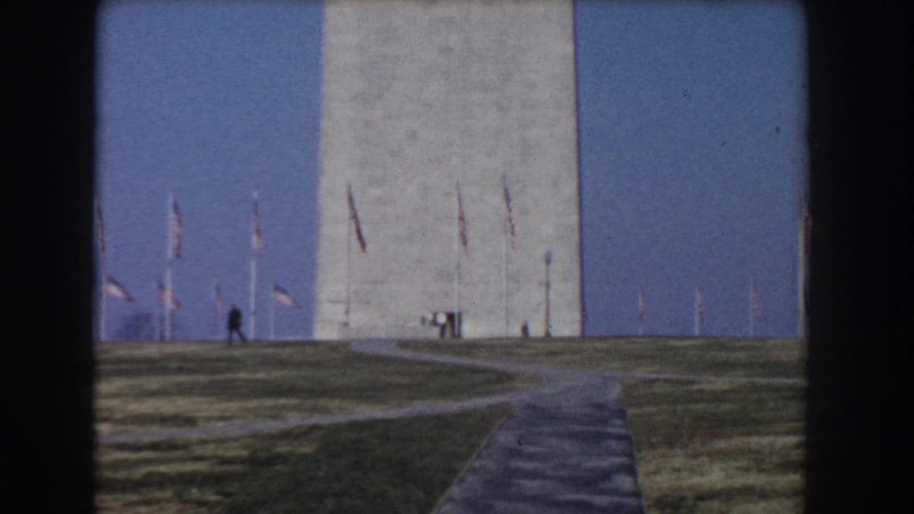 1960 8mm view of the Washington Monument in Washington, DC, seen beyond a path and lawn with American flags nearby.