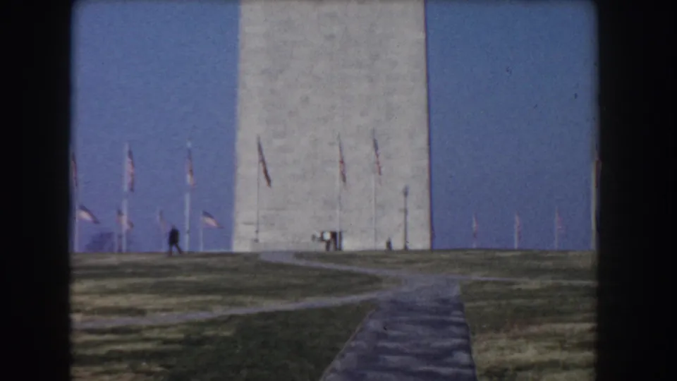 1960 8mm view of the Washington Monument in Washington, DC, seen beyond a path and lawn with American flags nearby.