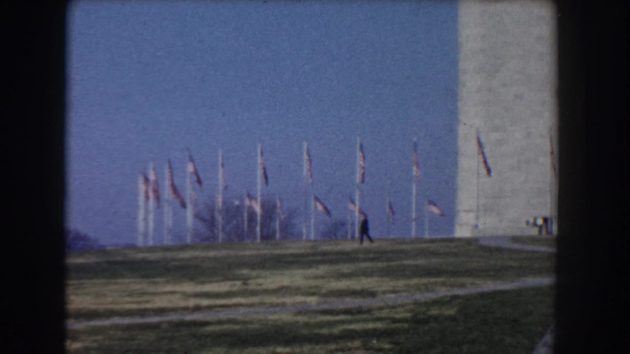 1960 Washington, DC home-movie frame showing American flags on poles near the Washington Monument with open grass in front.