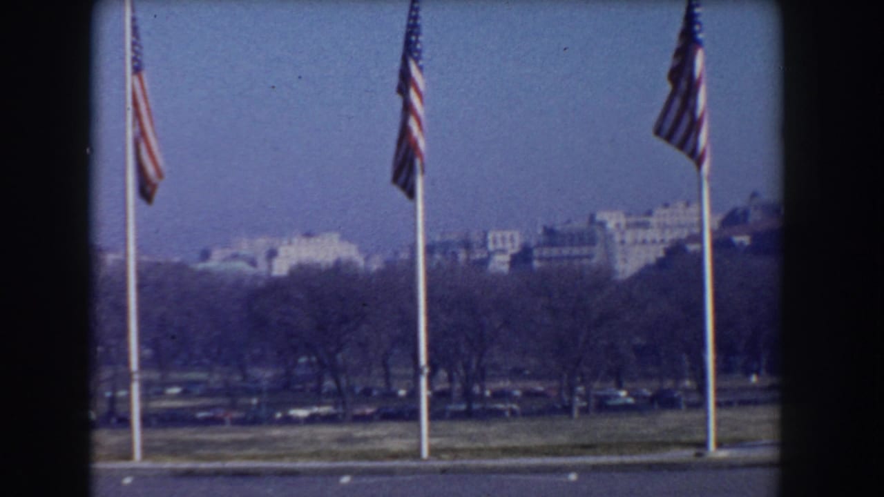 1960 Washington, D.C. home-movie frame showing American flags on tall poles near the National Mall with distant buildings beyond bare trees.