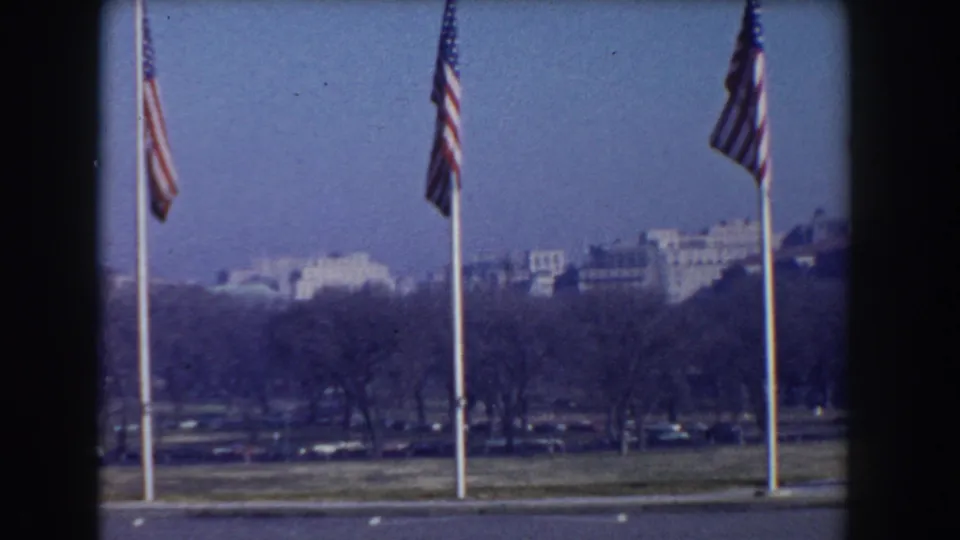 1960 Washington, D.C. home-movie frame showing American flags on tall poles near the National Mall with distant buildings beyond bare trees.