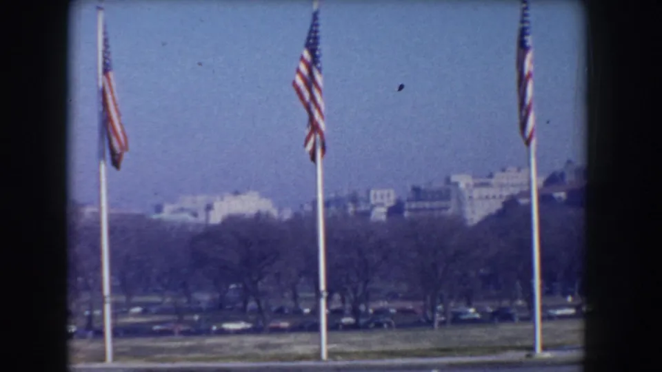 American flags flutter on tall poles in a 1960 Washington, D.C. view with the National Mall landscape and distant civic buildings visible.