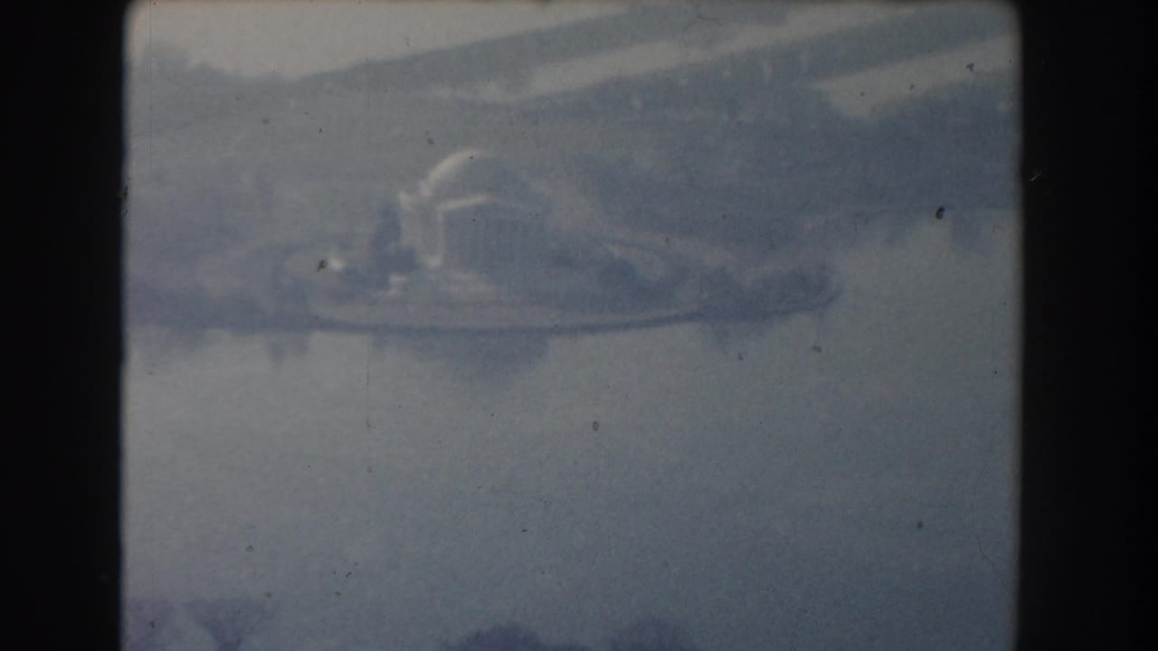 Hazy 1960 Washington, DC aerial frame showing water, shoreline, and distant urban forms through soft blue 8mm film texture.