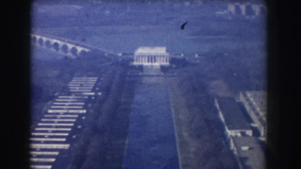 Blue-tinted 1960 Washington, DC aerial frame showing the Lincoln Memorial, Reflecting Pool, arched bridge, water, and surrounding grounds.