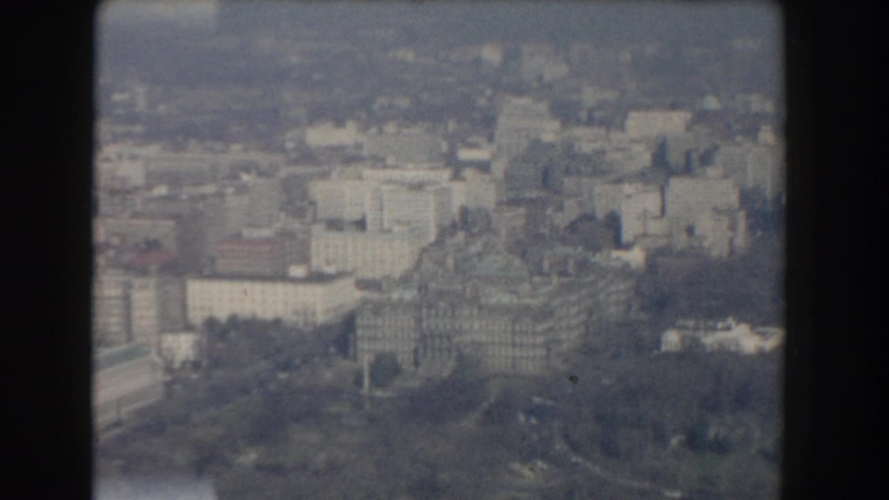 Soft aerial view of city buildings, parkland, and a castle-like stone structure.
