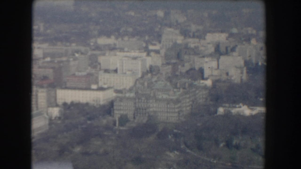 1960 Washington, DC aerial frame with institutional-looking buildings, tree-lined parkland, and hazy urban blocks beyond.