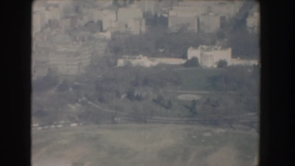 1960 Washington, DC aerial frame with open green space, curving roads, scattered trees, and institutional buildings in the distance.