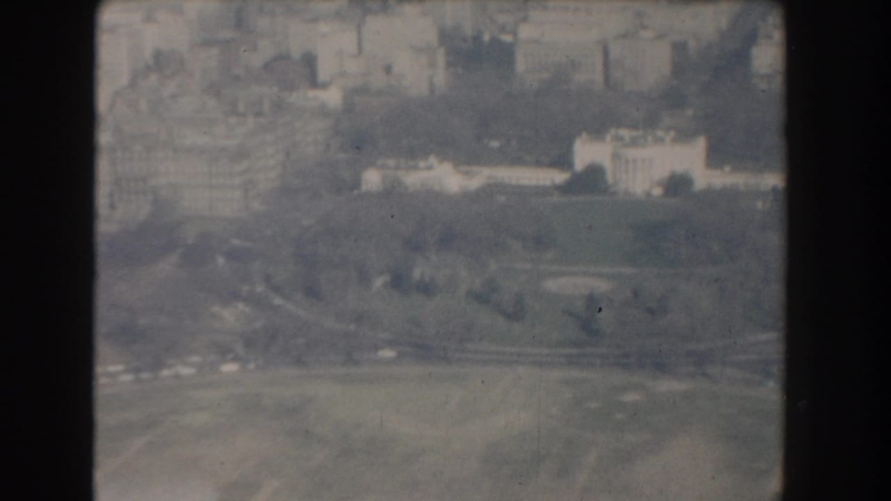 1960 8mm aerial frame looking across Washington DC toward the White House, with lawns, roads, and city buildings softened by haze.