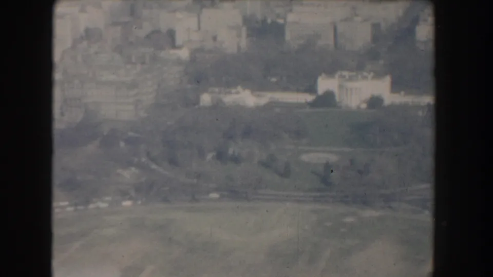 1960 8mm aerial frame looking across Washington DC toward the White House, with lawns, roads, and city buildings softened by haze.