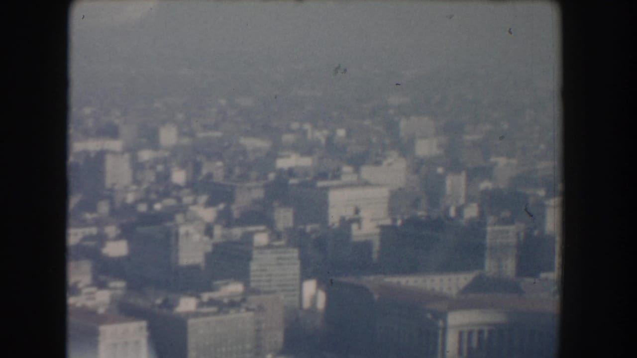 Hazy high-angle view of a city skyline with blocky buildings and dark film borders.