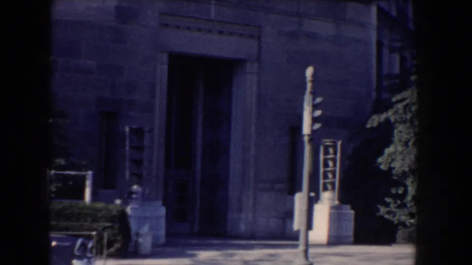 1960 Washington DC home-movie view of a gray stone building entrance, traffic signal pole, shrubs, and a tree at right.