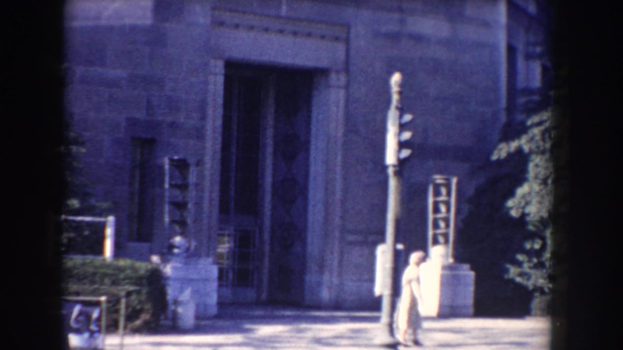 1960 Washington, DC home-movie frame showing a stone FBI building entrance, traffic signal, sidewalk, and pedestrians.