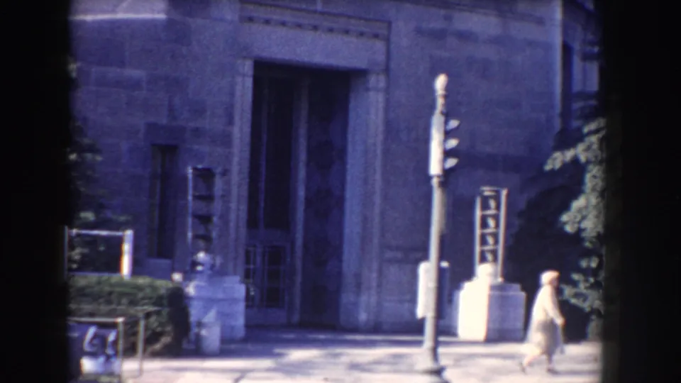 1960 Washington, DC frame with a woman walking past the FBI building entrance and traffic signal.