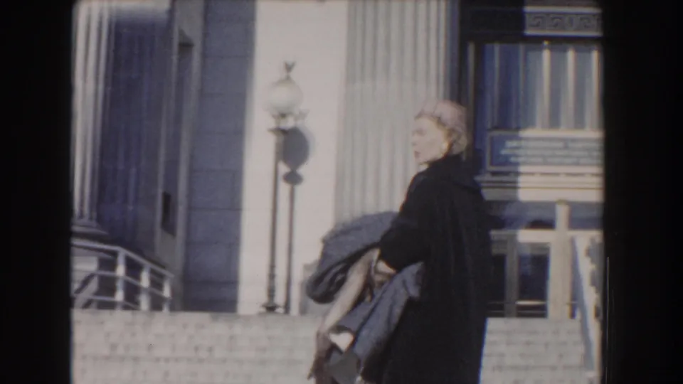 1960 Washington, DC frame with a woman in a dark coat and hat near stone steps, columns, lamps, and the building entrance.