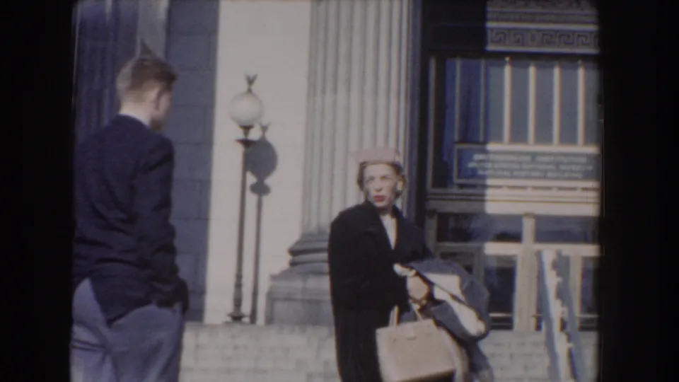 1960 Washington, DC home-movie frame showing a man and a woman by stone steps, a column, globe lamp, entrance doors, and signage.