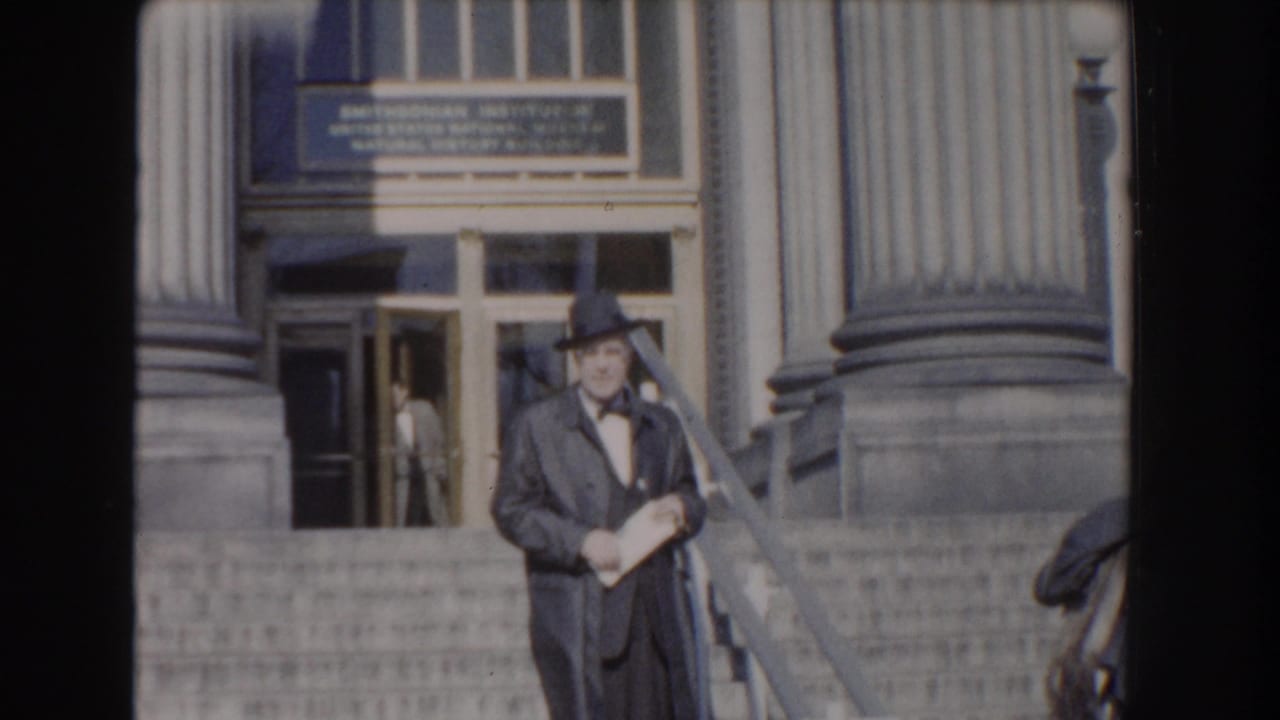 Man in dark coat and hat holds papers on stone steps outside a columned entrance.