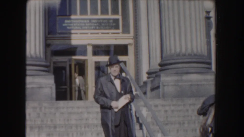 In Washington DC archival footage, a formally dressed man stands on broad steps near columns and entrance doors, holding papers.