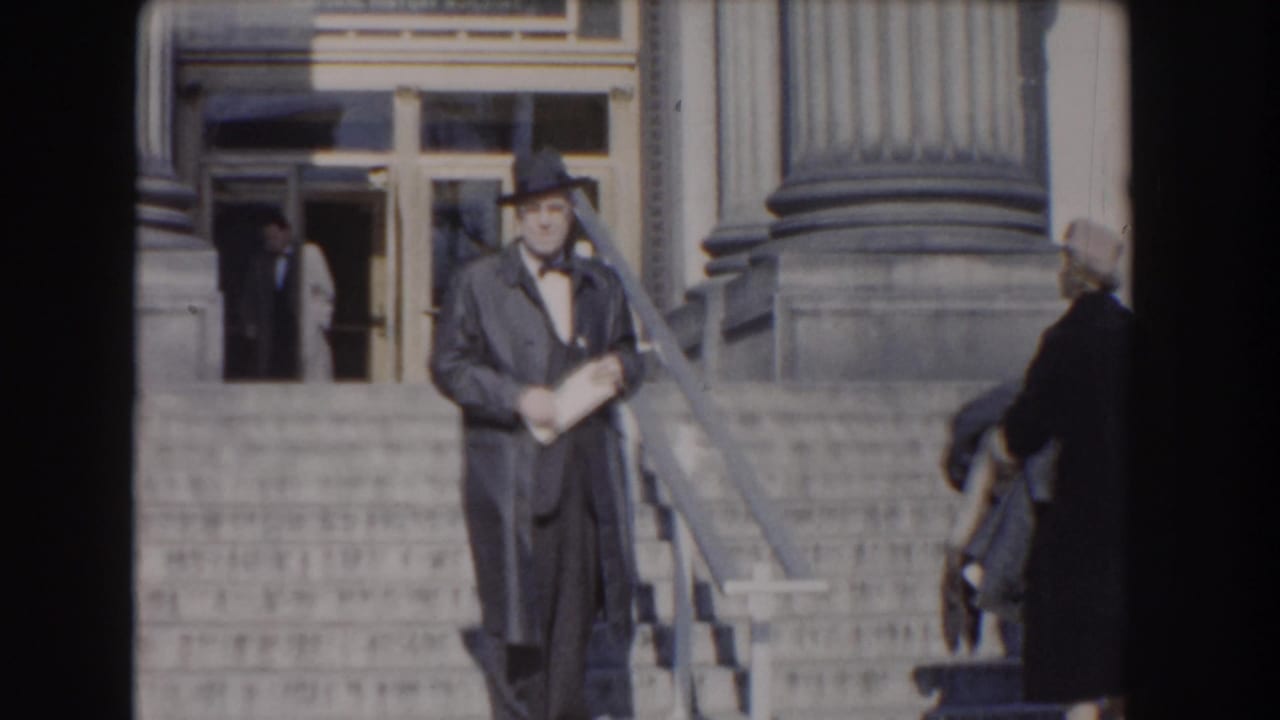 Man with papers faces the camera on the steps while another person appears near the doorway.