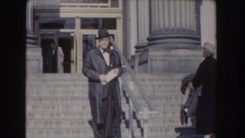 The suited man remains on the steps of the Washington DC building, with columns, entrance doors, and another adult near the doorway.
