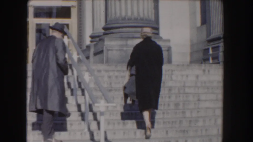 Two adults in period outerwear walk up the broad Washington DC steps toward the columned entrance, seen from a handheld home-movie view.