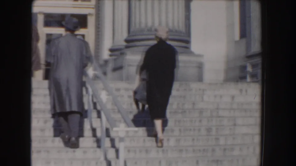 From behind, two adults continue up the wide stone staircase toward the columns and doors of the Washington DC institutional building.