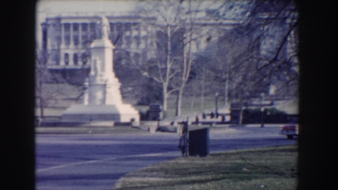 White monument and distant classical building seen across a park road.