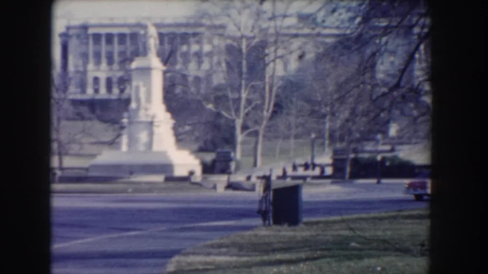 A white monument stands near a roadway with a classical government-style building behind it in Washington, DC.