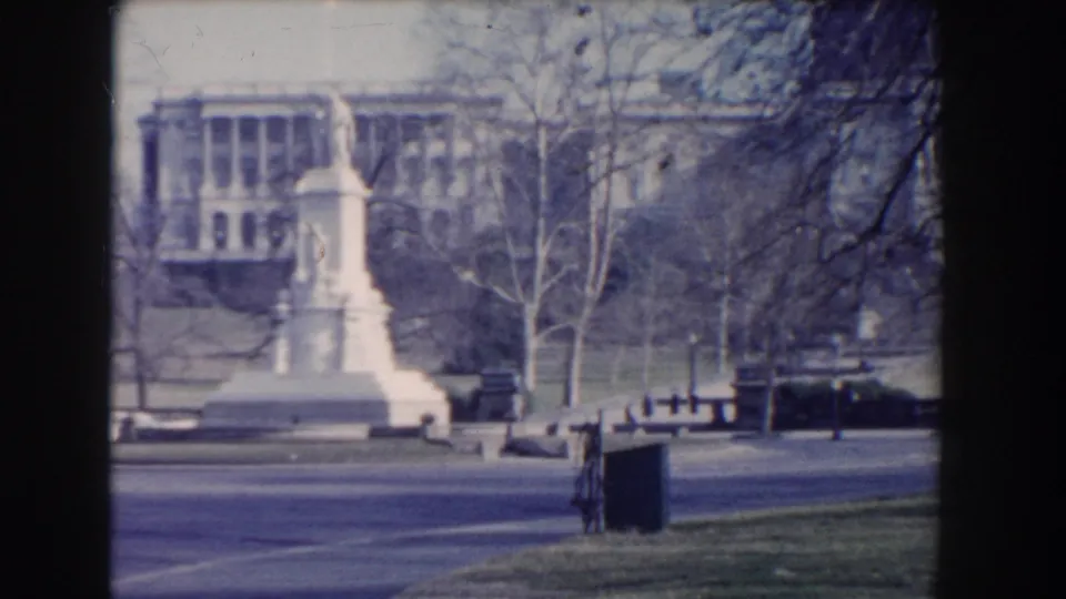 Washington, DC park view showing a white monument beside a road, with bare trees and a distant civic building.