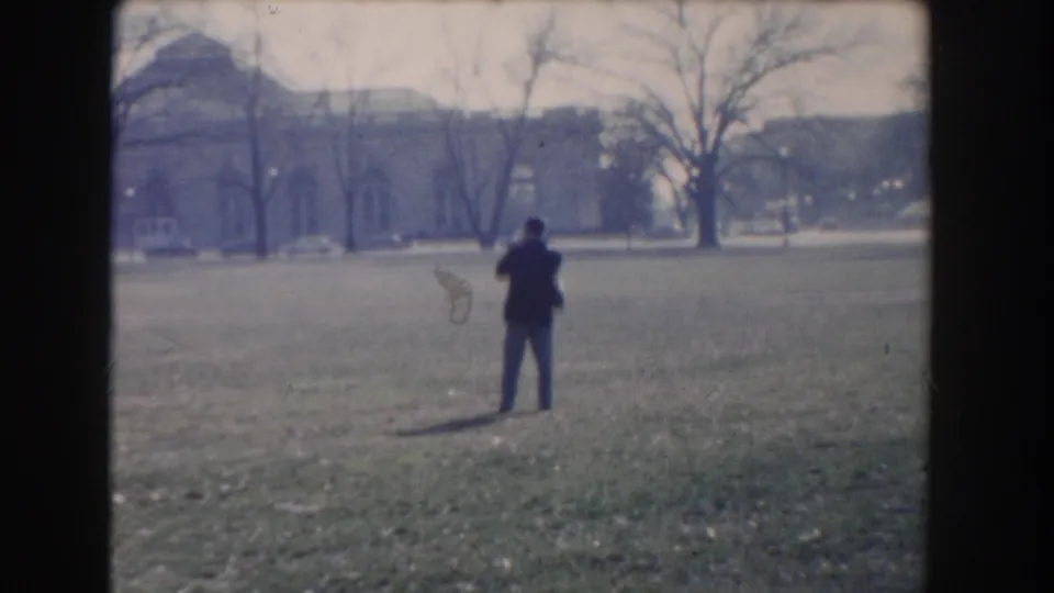 A man in a dark coat stands in a Washington, DC park filming the landscape with bare trees and distant buildings behind him.