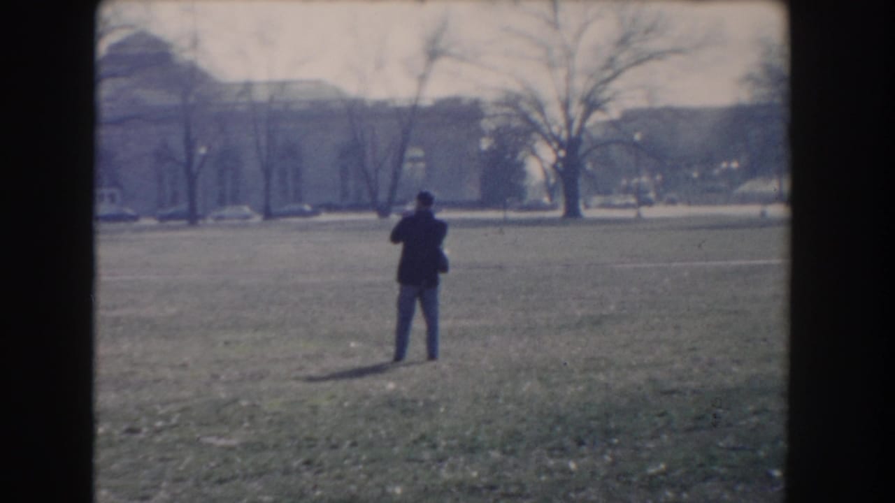 Man standing in a grassy park with bare trees and buildings in the distance.