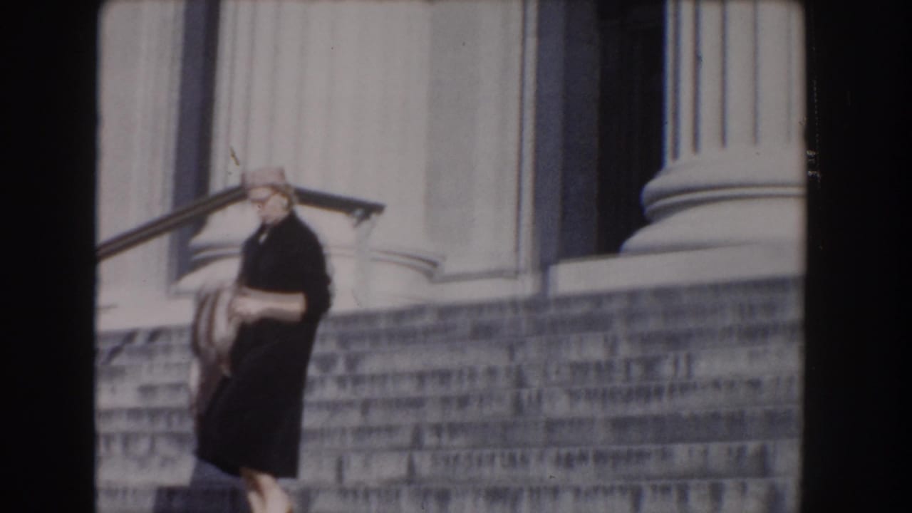 Woman in a dark coat and hat walks down stone steps beside large columns.