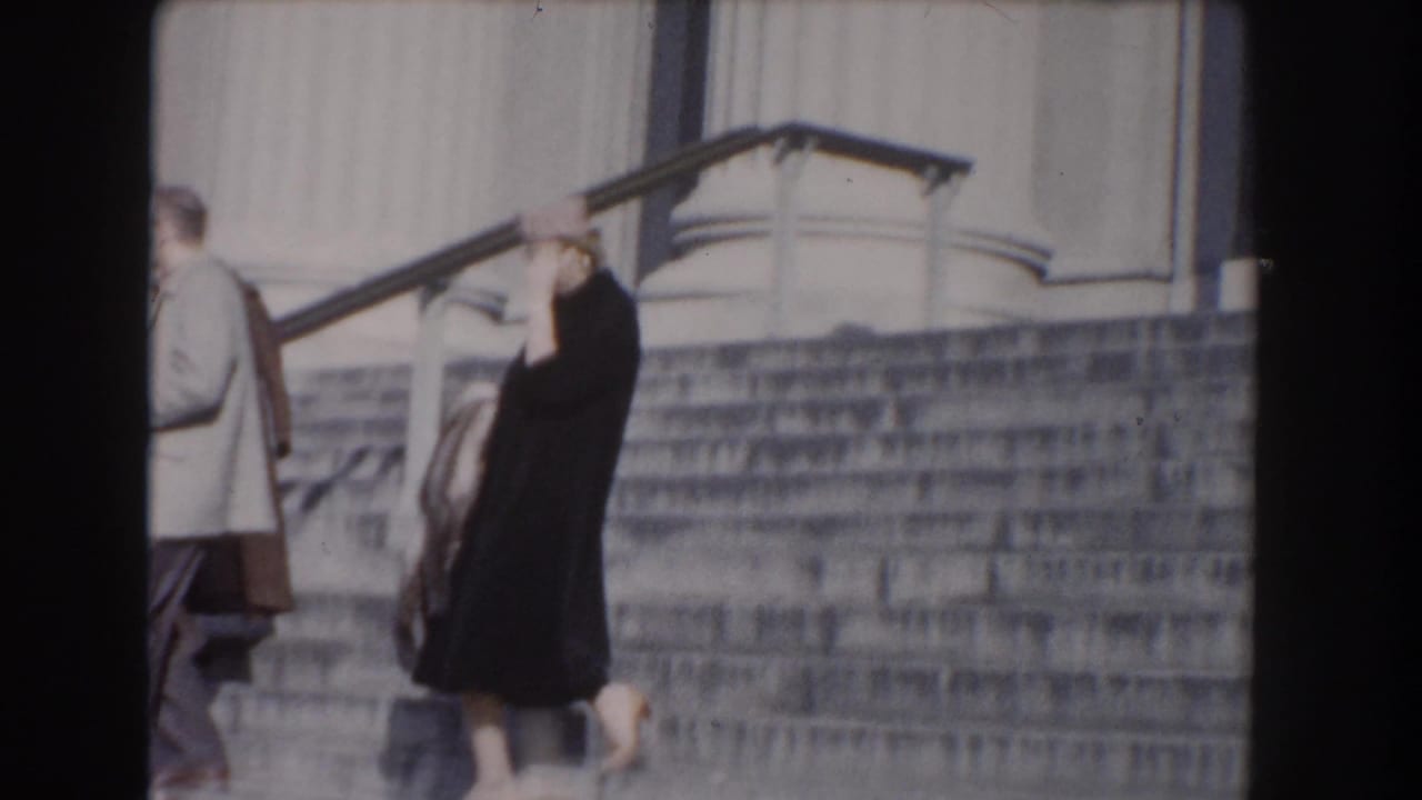A woman in a dark coat and hat passes a man in a light jacket on the steps of a Washington, DC neoclassical building.
