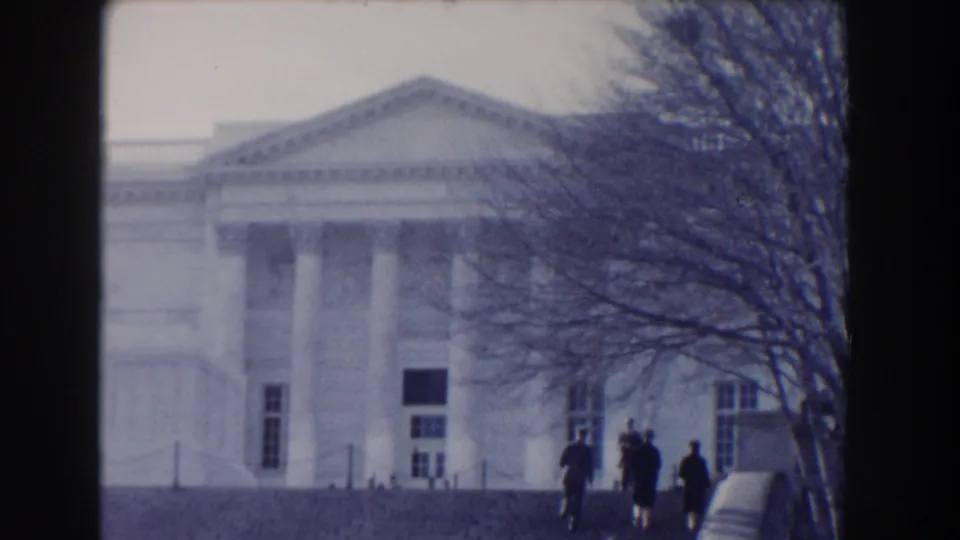 Wide 1960 Washington DC home-movie frame showing a white neoclassical building, bare trees, and several distant pedestrians.