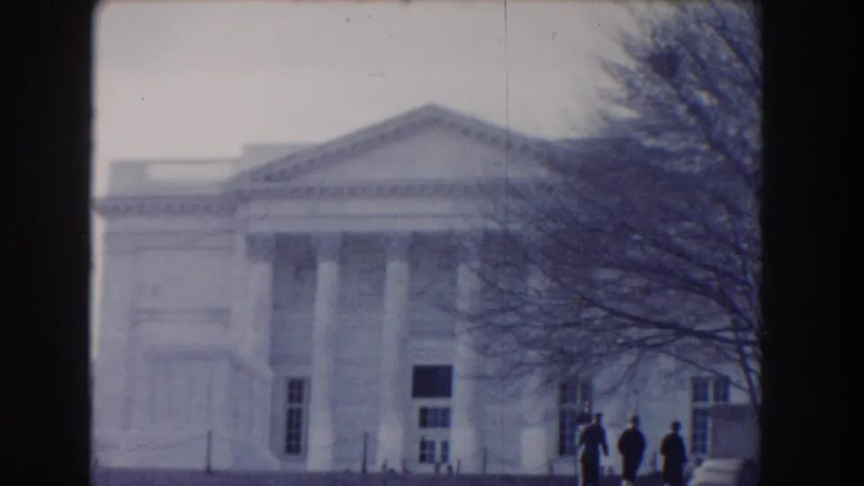 Soft 8mm frame of a Washington DC neoclassical facade with a pediment, tree branches, and distant people in period clothing.