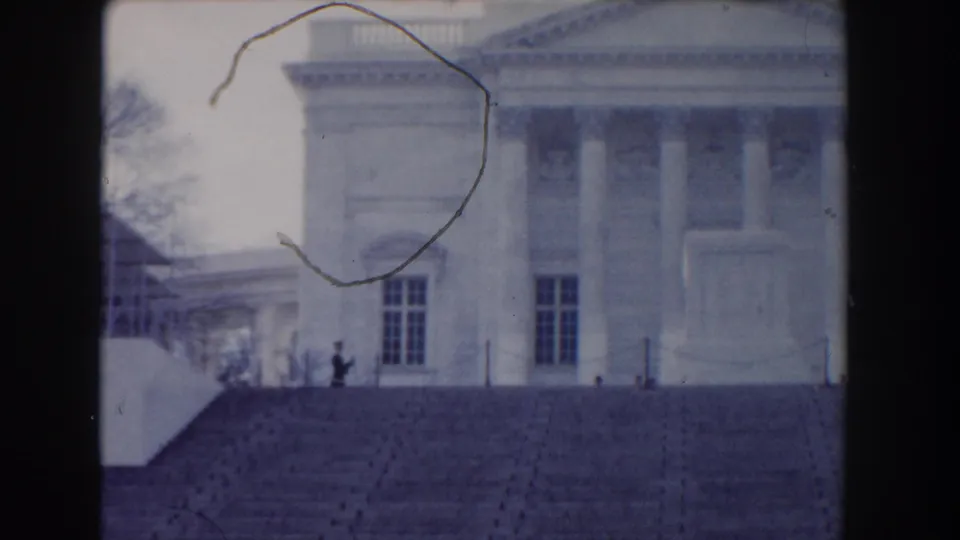 1960 home-movie frame with visible film scratches over a distant guard and the pale facade of the memorial building.