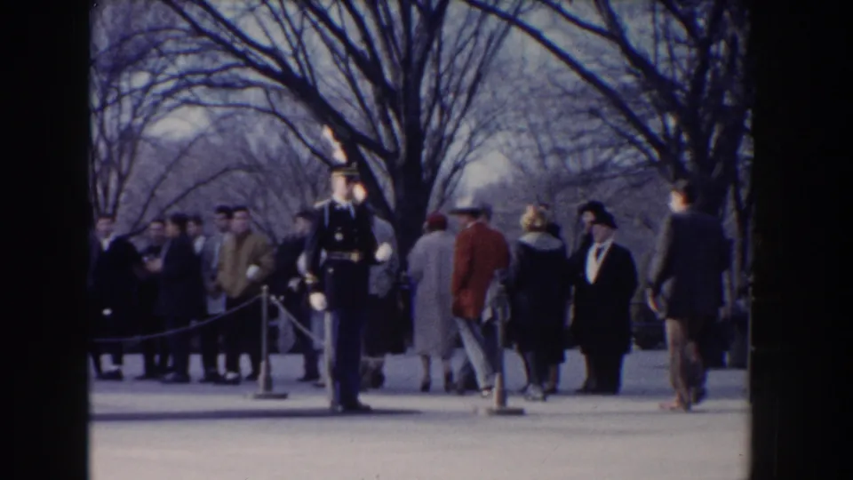 1960 Washington DC frame with a uniformed rifle-bearing guard standing before civilians gathered behind roped barriers.