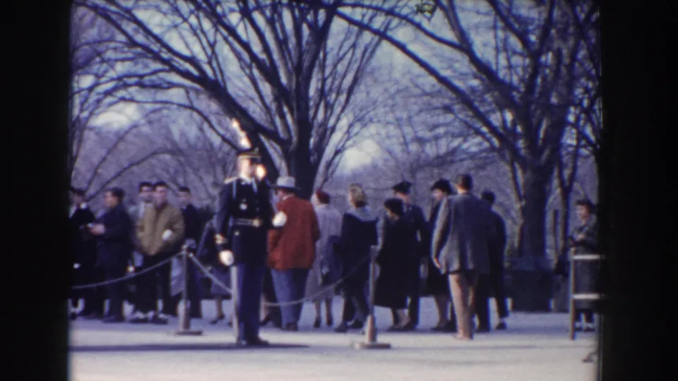 1960 Washington DC home-movie frame showing a guard with a rifle near formally dressed civilians and roped visitor lines.