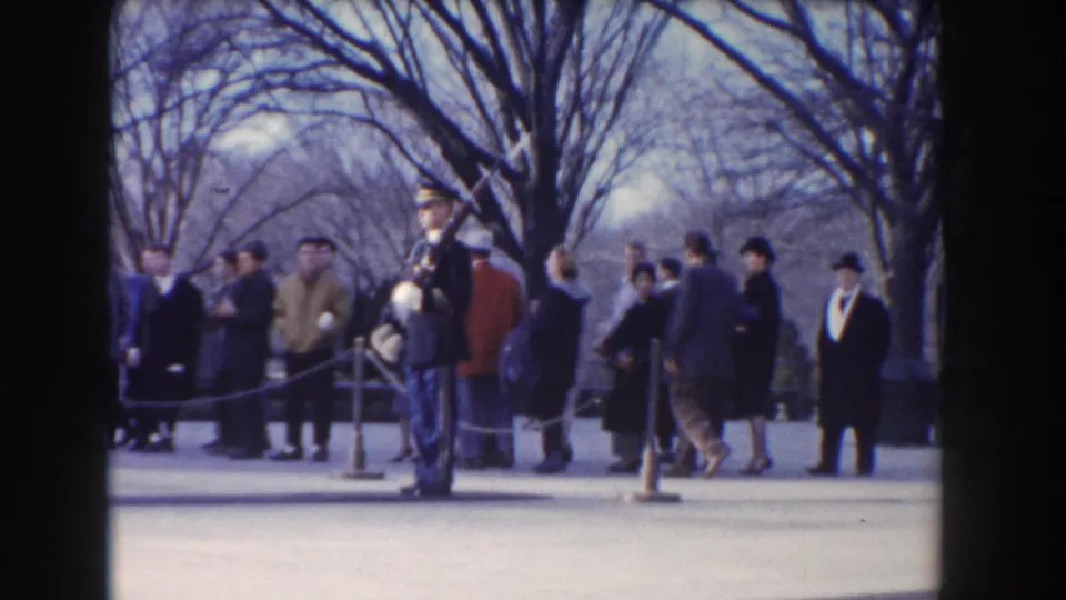 1960 Washington DC frame showing a uniformed armed guard standing near civilians in coats beside roped barriers and leafless trees.