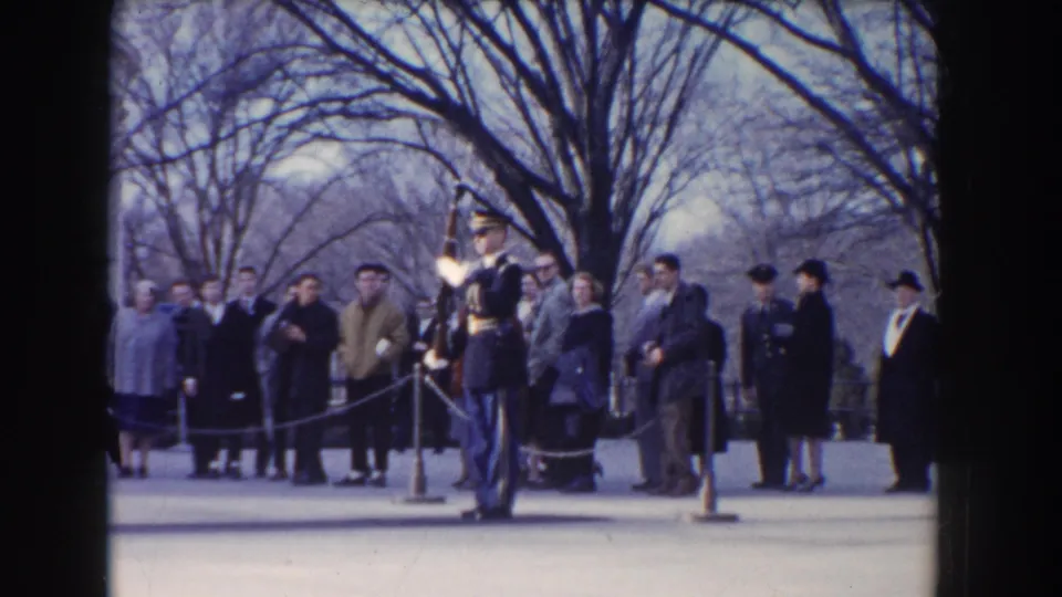 In 1960 Washington, DC footage, a ceremonial guard with rifle stands at attention while visitors watch from behind a rope barrier.