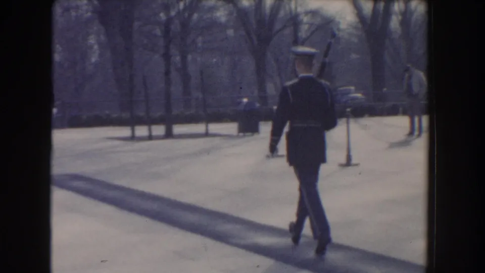 A ceremonial guard in Washington, DC walks away from the camera across a paved park path, casting a long shadow.