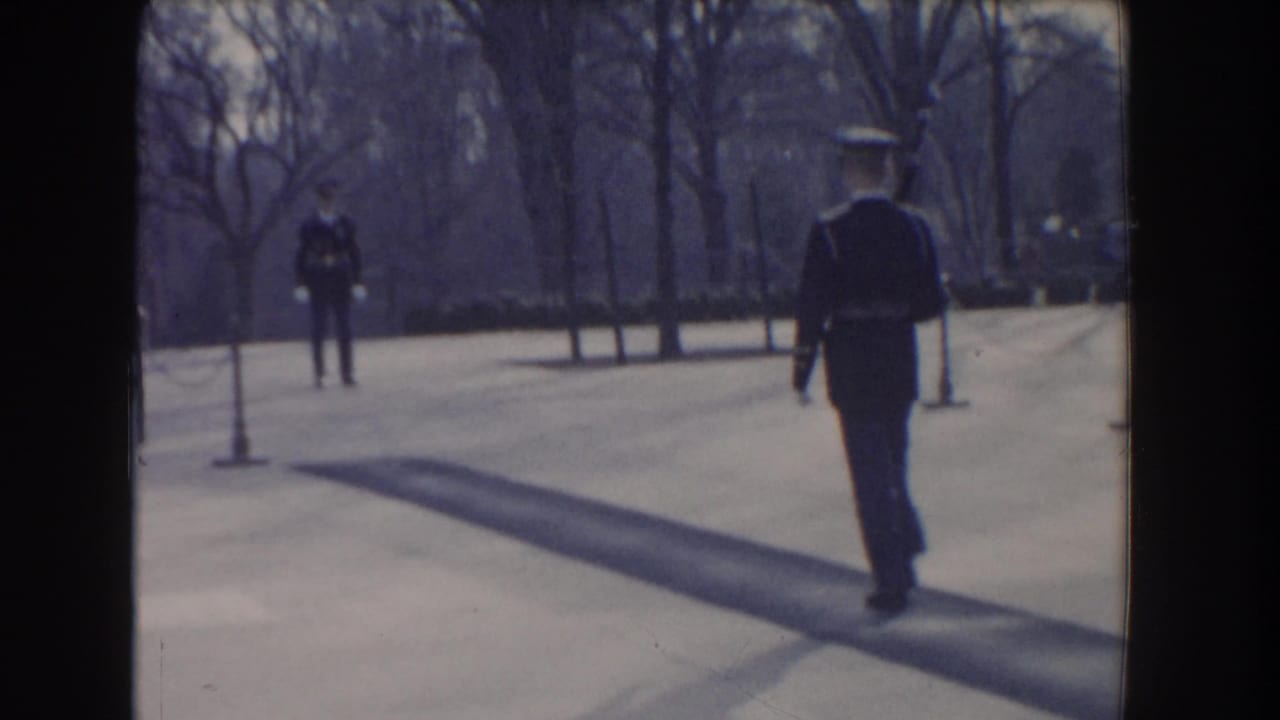 In Washington, DC, a uniformed guard walks across a paved monument plaza, with another figure and bare trees in the distance.