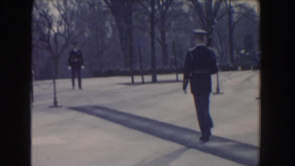 In Washington, DC, a uniformed guard walks across a paved monument plaza, with another figure and bare trees in the distance.